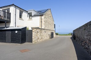 A residential street with a white house, a stone wall, and a clear view of the sea.