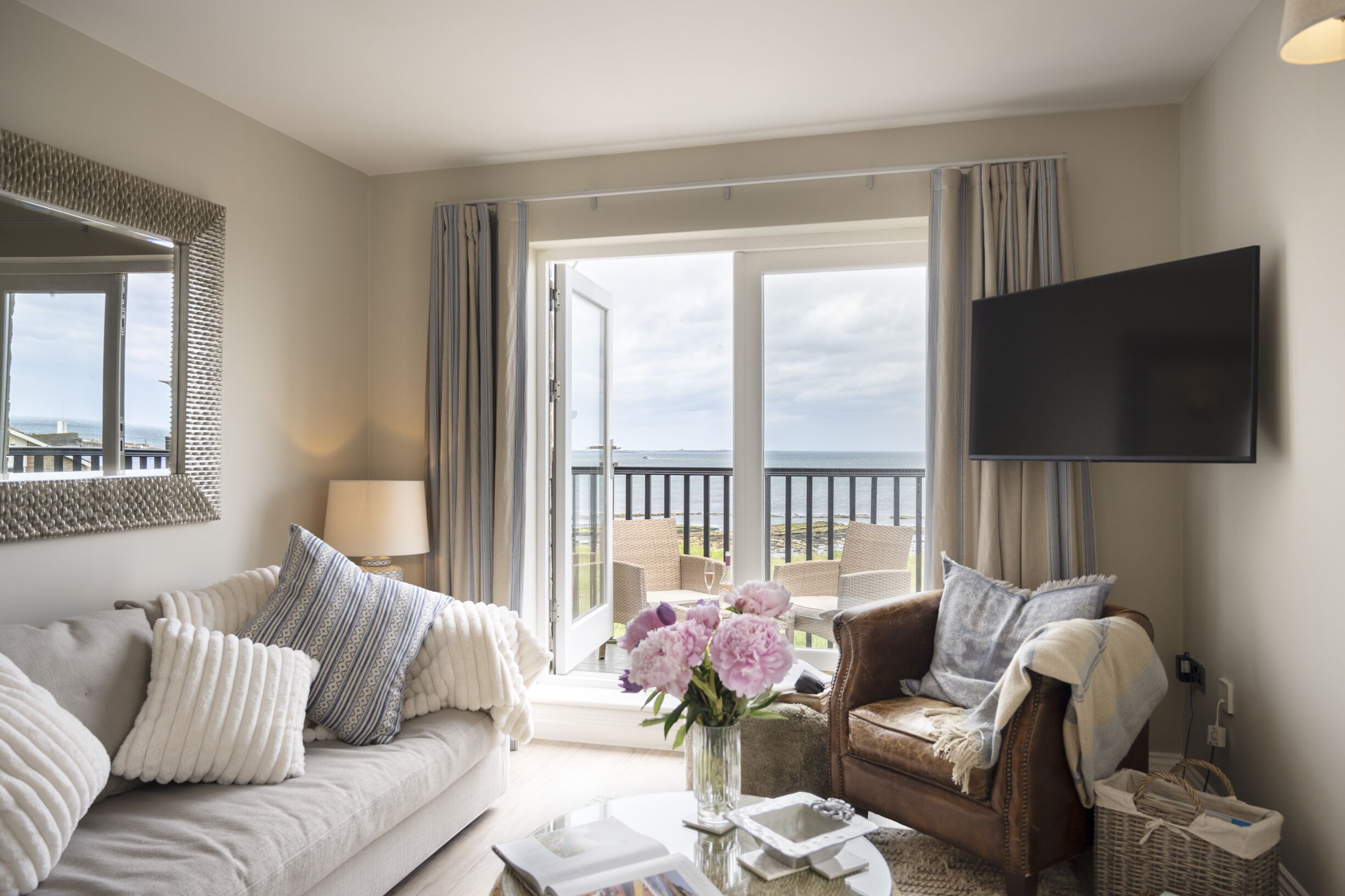 Un salon spacieux comprenant un canapé, un fauteuil et une table basse. Un téléviseur est fixé au mur et, par la porte ouverte, on peut admirer la vue sur la mer.
