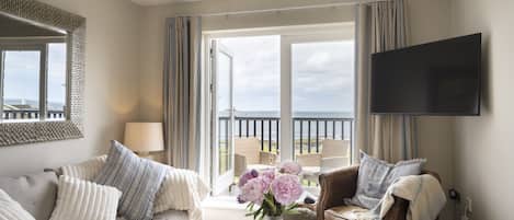 A spacious living room with a sofa, armchair, and a coffee table. There is a television mounted on the wall and a view of the sea through the open door.