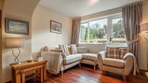 Un salon chaleureux avec un plancher en bois, un canapé, un fauteuil et une table. Il y a deux fenêtres avec des rideaux, une lampe et des tableaux encadrés accrochés au mur.