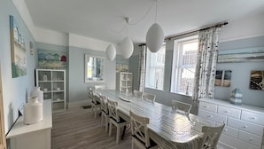 A dining area with a long table, white chairs, and pendant lights. There are windows with curtains, a white cabinet with decorative items, and wall art.