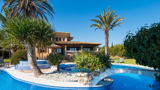 Une piscine avec une maison en arrière-plan, entourée de palmiers et surplombée par un ciel bleu clair.