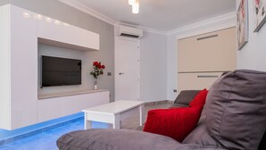 Una sala de estar moderna con un televisor de pantalla plana montado en la pared, una mesa de centro blanca, un sofá gris con cojines rojos y un mueble beige.