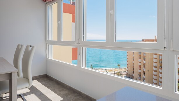 Una habitación con un amplio ventanal que da a la playa y edificios. Hay una mesa y dos sillas.