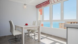 Un comedor moderno con una mesa y sillas blancas, una manzana roja sobre la mesa, y vistas al mar a través de amplias ventanas.