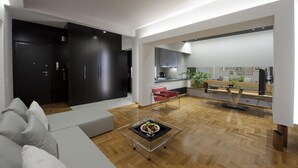 Una sala de estar moderna con un sofá, una mesa de centro con fruta, una silla roja y una cocina al fondo.