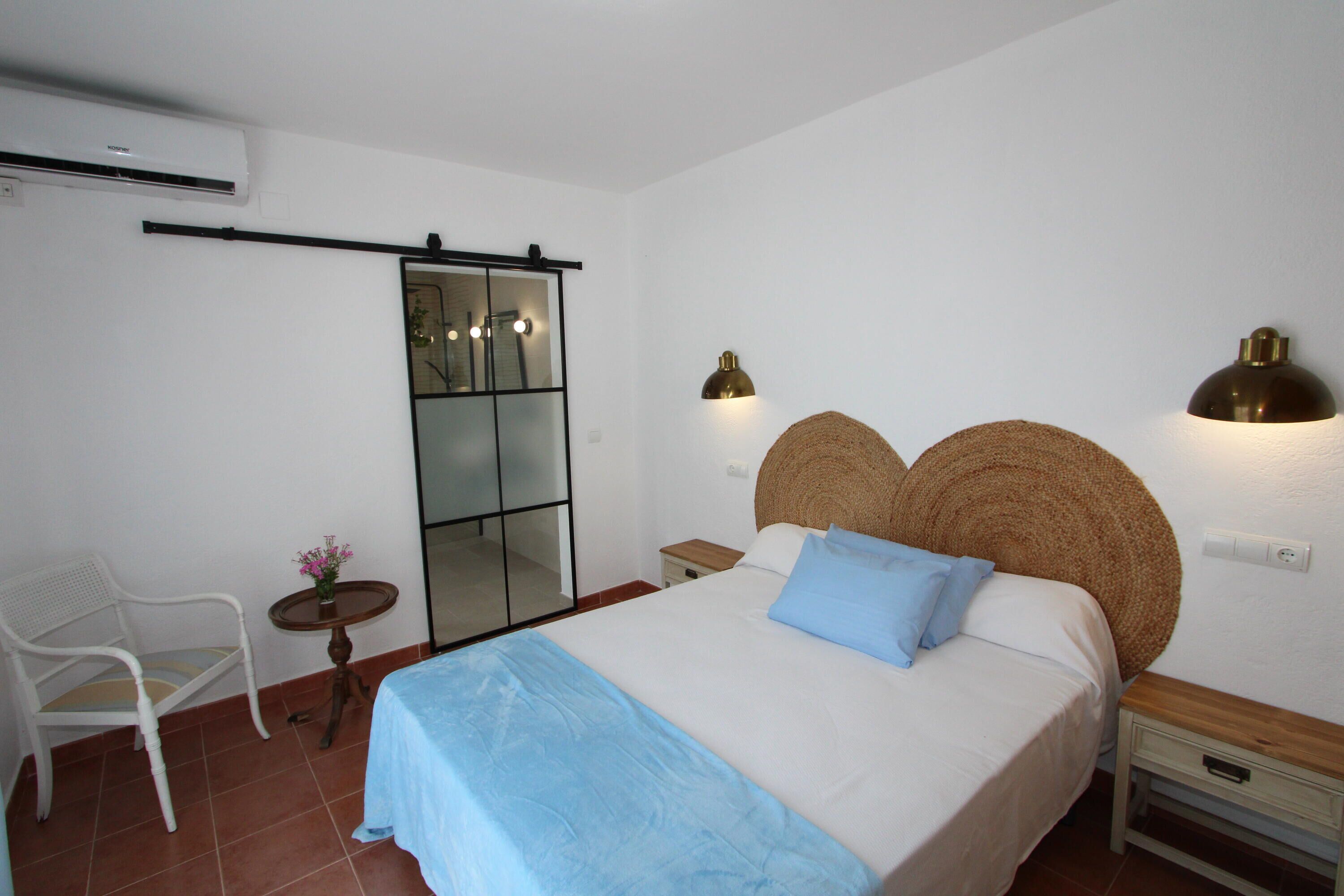 A bedroom with a bed, a chair, a small table with a vase of flowers, and a mirror.