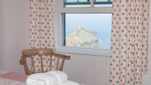 A bedroom with a bed, a chair, and a window with curtains. - Holiday cottage with beautiful views over the Cornish sea (St Ives)