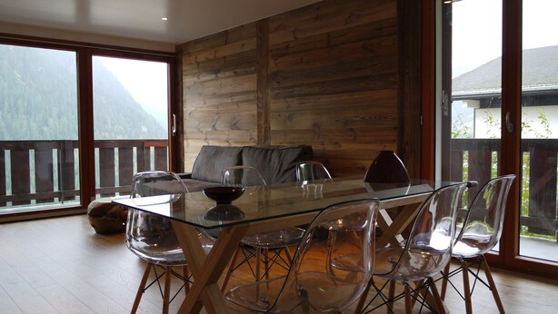 A dining area with a glass table and clear chairs, a sofa, and a mountain view through large windows. - Chatel Centre - 50 M to Ski Lift (Châtel)
