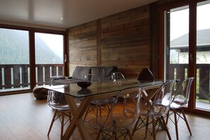 Un coin repas avec une table en verre et des chaises transparentes, un canapé et une vue sur les montagnes par de grandes fenêtres.