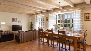 Un comedor con una mesa y sillas de madera, un sofá y un televisor en una sala de estar con piso de baldosas y vigas de madera en el techo.