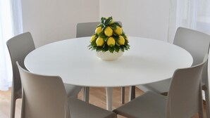 Ein runder, weißer Tisch mit floraler Dekoration, umgeben von vier grauen Stühlen, in einem Raum mit Holzboden und einem an der Wand befestigten Fernseher.