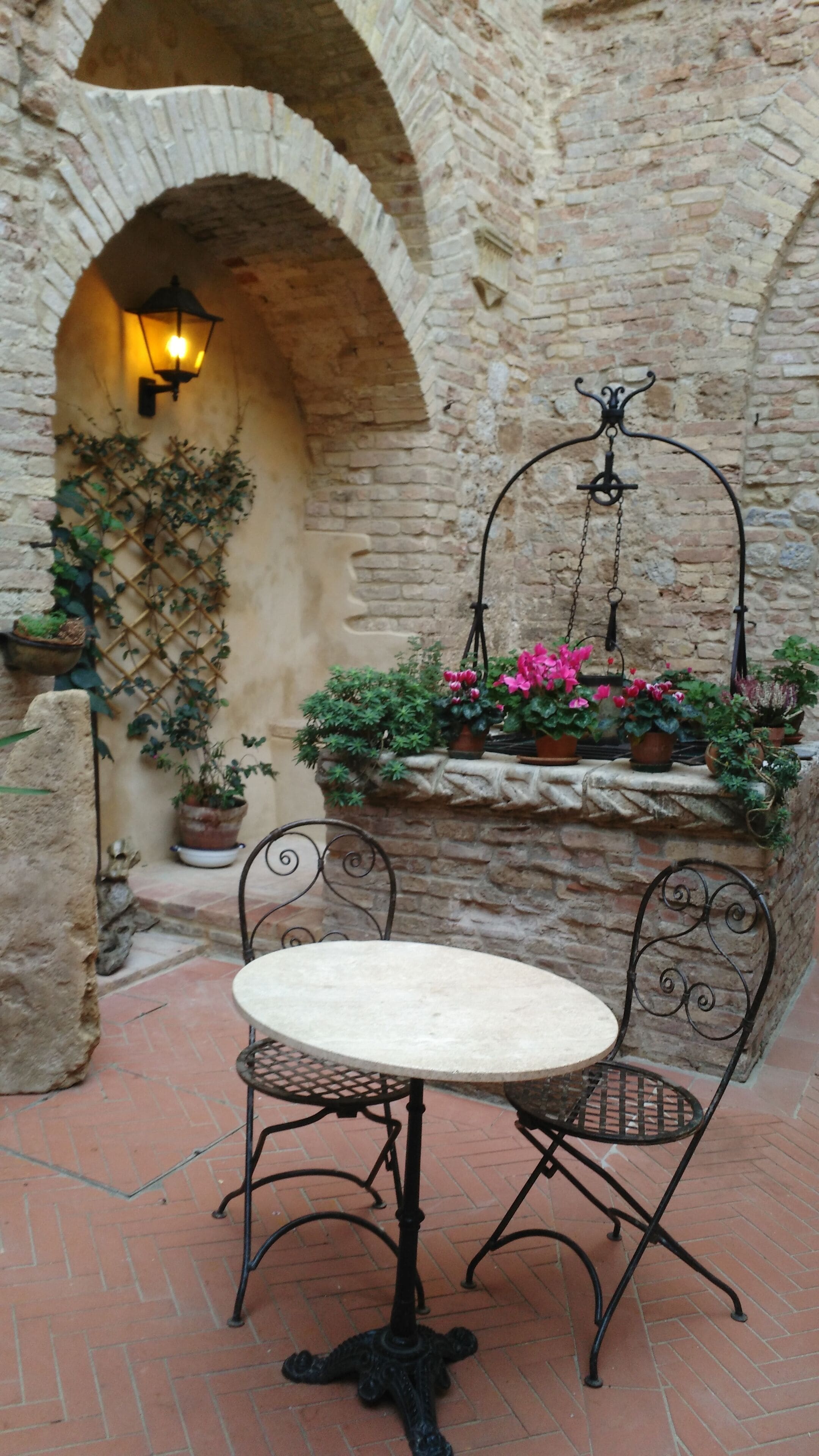 Un arco de piedra con un aplique colgante, una mesa redonda con sillas de metal y plantas en macetas sobre un suelo de ladrillo.
