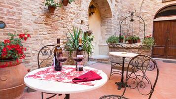 An outdoor patio with a round table, two chairs, and a wine bottle. There are potted plants and a stone wall in the background.