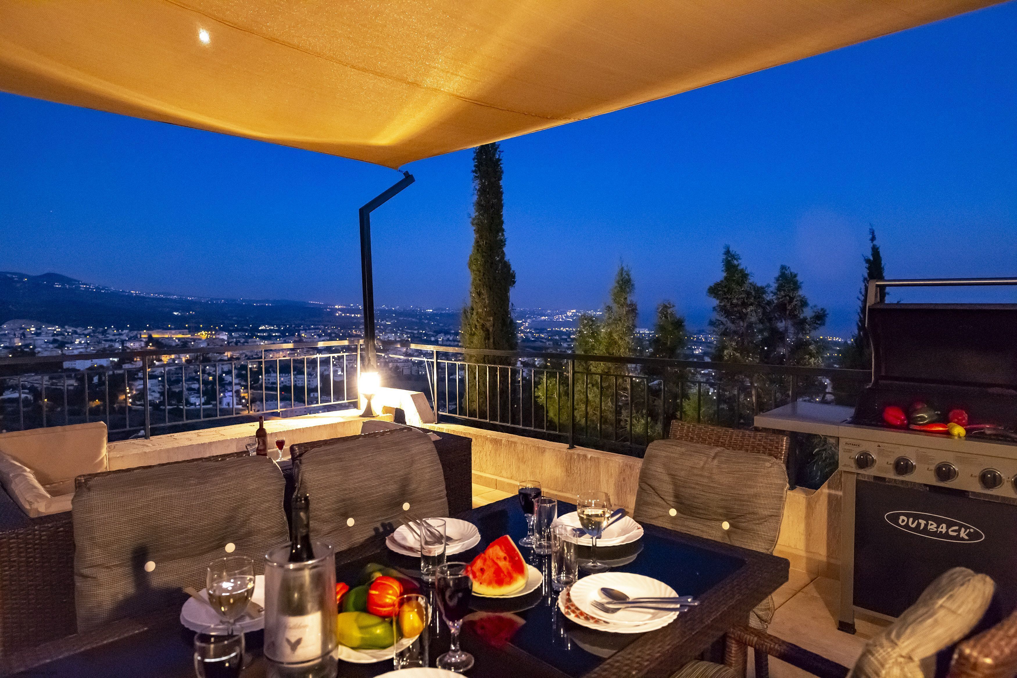 A rooftop dining area with a table set for two, a grill, and a view of the city at dusk.