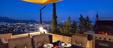 Un área de comedor en la azotea con una mesa dispuesta para dos personas, una parrilla y vistas de la ciudad al atardecer.