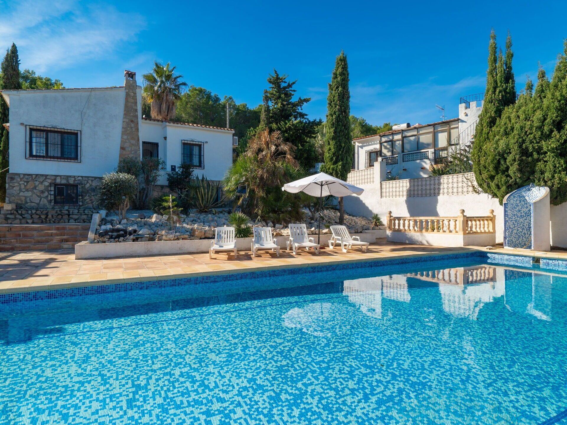 A swimming pool with tiled blue edges, surrounded by white lounge chairs and a white umbrella.