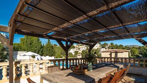 Um pátio externo com mesa e cadeiras de madeira, uma pérgola acima e vista para uma piscina e edifícios ao fundo.