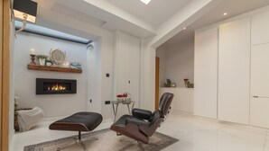 A modern living room with a fireplace, a black leather chair, a small side table with decorative items, and a white wardrobe.