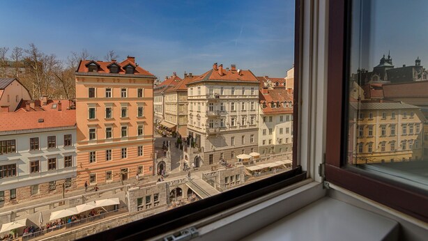 A view of a historic European town from a window, featuring multi-story buildings with red-tiled roofs and a central square with a staircase.