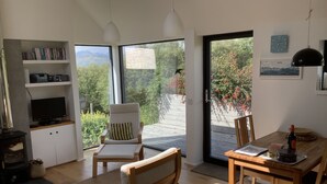 Ein modernes Wohnzimmer mit Fernseher, Bücherregalen, Kamin und Blick auf einen Garten und Berge durch große Fenster.