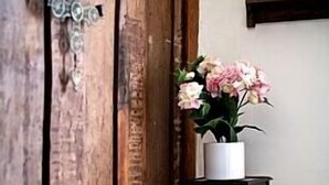 Una puerta de madera con un tirador de metal decorativo, un jarrón blanco con flores rosas sobre un taburete pequeño, y un espejo en la pared.