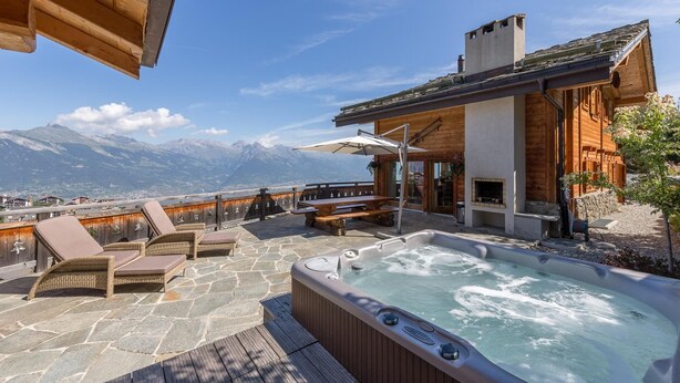 A hot tub on a stone deck with lounge chairs and a wooden building with a fireplace. - Beautiful chalet in the Swiss Alps with stunning views and private hot tub  (Haute Nendaz)