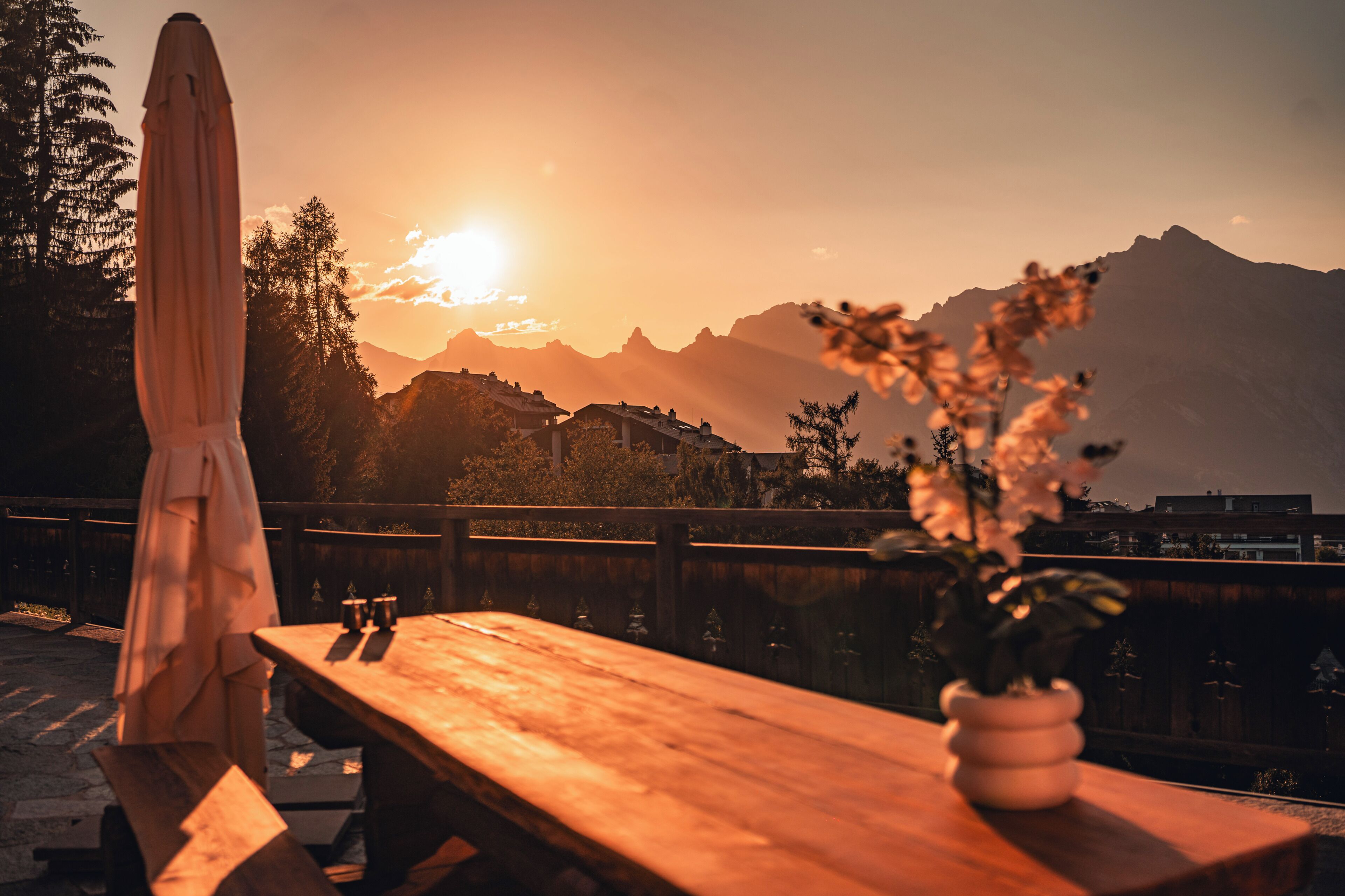 A wooden table with a vase of flowers, an umbrella, and a view of mountains during sunset.