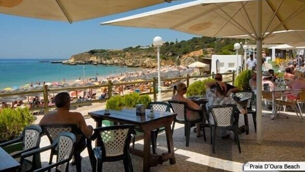 Un espace extérieur aménag é avec des tables et des chaises, donnant sur une plage parsemée de parasols et fréquentée par des gens.