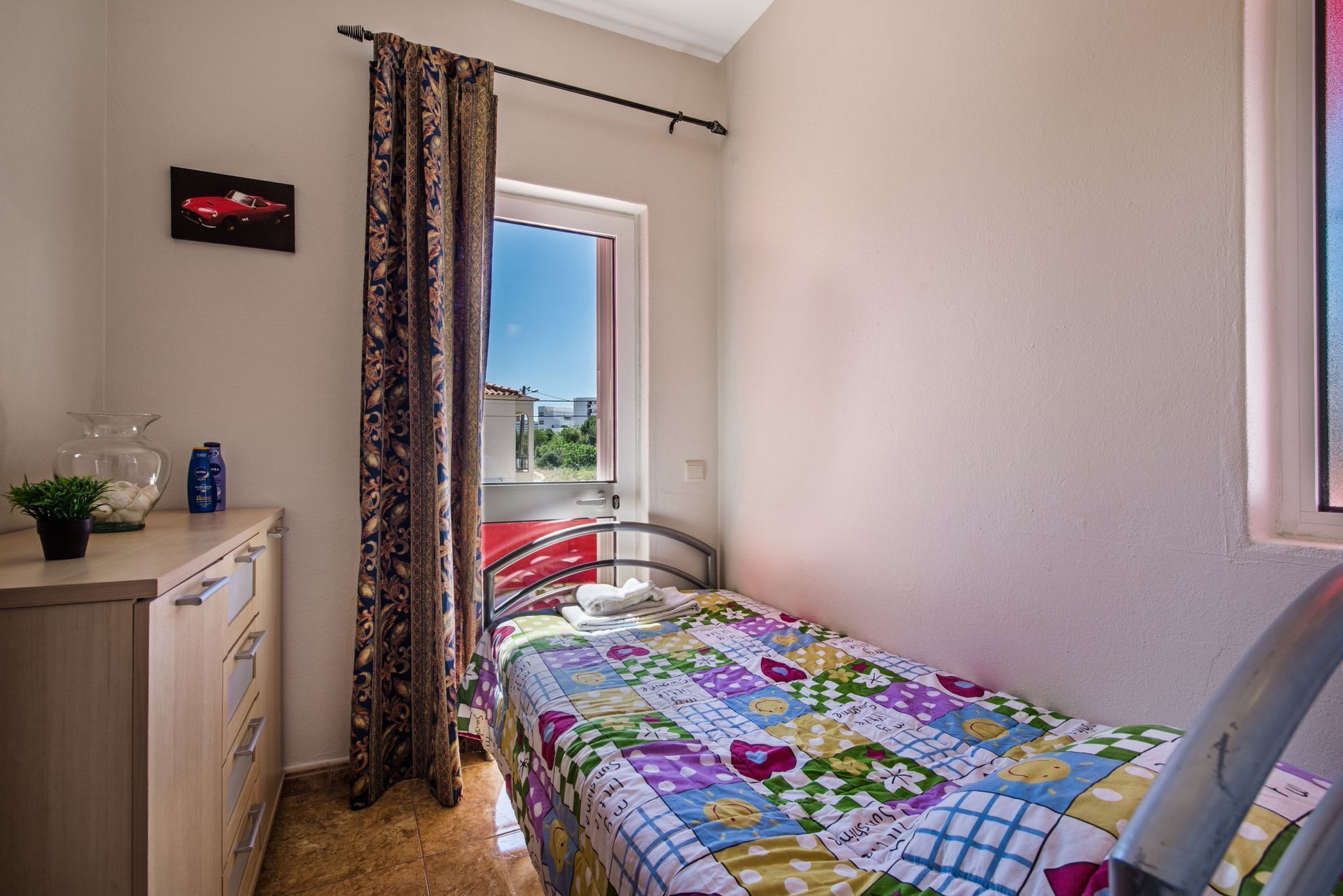 A bedroom with a bed, a window with curtains, a small table with a plant, and a wall-mounted picture.
