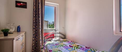 A bedroom with a bed, a window with curtains, a small table with a plant, and a wall-mounted picture.