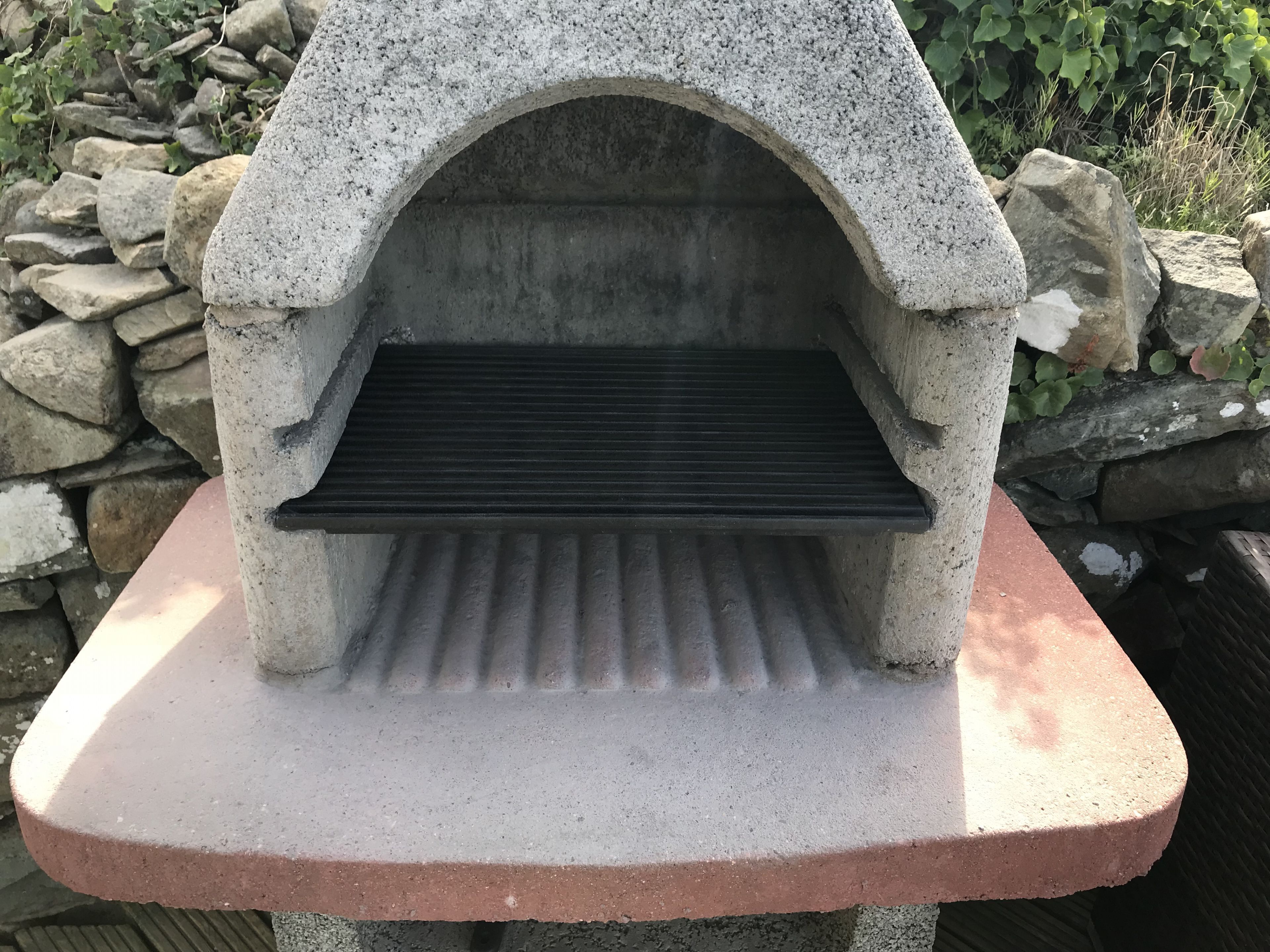 Un horno de pizza de piedra con una rejilla de metal, sobre una base de ladrillo, rodeado de un muro de piedra y vegetación.