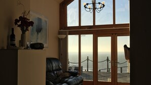 A spacious living room with a large bay window overlooking the sea, a leather sofa, a chandelier, and a painting on the wall. - 5 Star Luxury barn conversion overlooking the sea in North Wales (sleeps 2) (Barmouth)