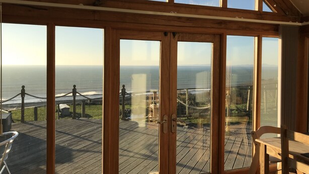 A room with large windows offering a view of the ocean and a deck area with a table and chairs. - 5 Star Luxury barn conversion overlooking the sea in North Wales (sleeps 2) (Barmouth)