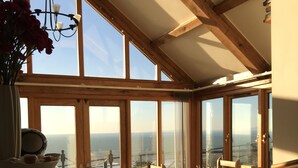 A sunlit room with a wooden table and chairs, large windows with a view of the sea, and a vase with flowers on a table. - 5 Star Luxury barn conversion overlooking the sea in North Wales (sleeps 2) (Barmouth)