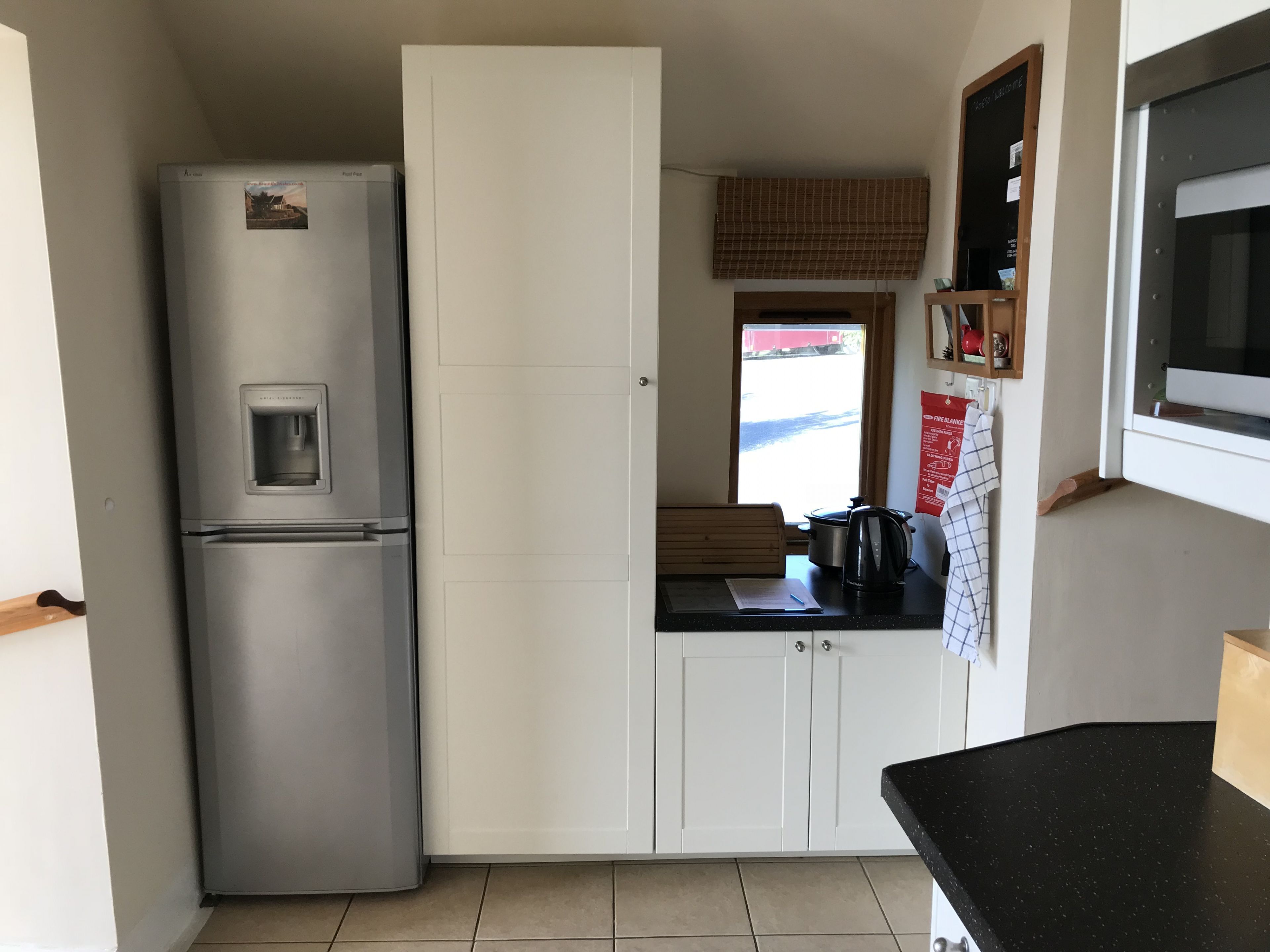 Ett kompakt kök med ett kylskåp i rostfritt stål, vita skåp, en svart bänkskiva och ett fönster med utsikt över gatan.