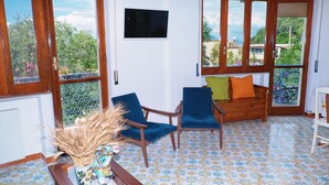 Una habitación con suelo de baldosas, dos sillas, un sofá, un televisor y una ventana con vistas a zonas verdes y edificios.