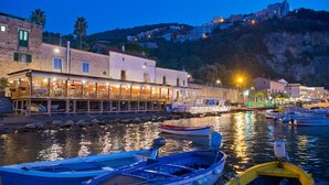 Un edificio frente al agua con un restaurante en la planta baja, barcos amarrados en primer plano y una colina con edificios al fondo.