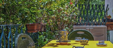 Mesa amarela posta com bules e xícaras azuis e amarelas, e uma bandeja, rodeada por plantas verdes e uma cerca de metal.