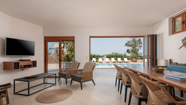 A modern living room with a flat-screen TV, a wooden dining table, wicker chairs, and a pool view. - Tsourlakis Residence, A Tranquil Family Oasis with Pool (Rethymno)