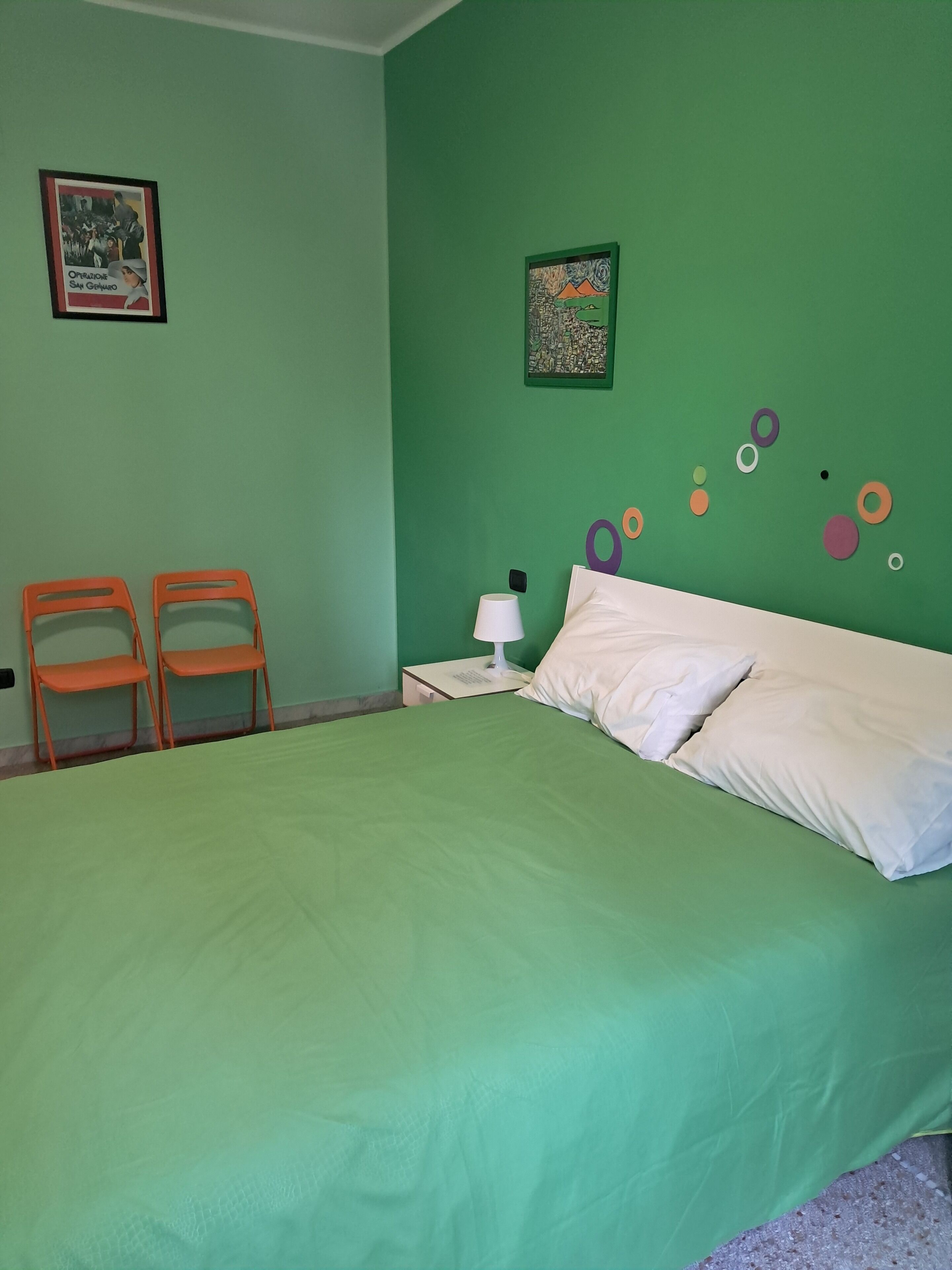 Una habitación con cama, dos sillas, una mesita de noche con lámpara y decoración en las paredes.