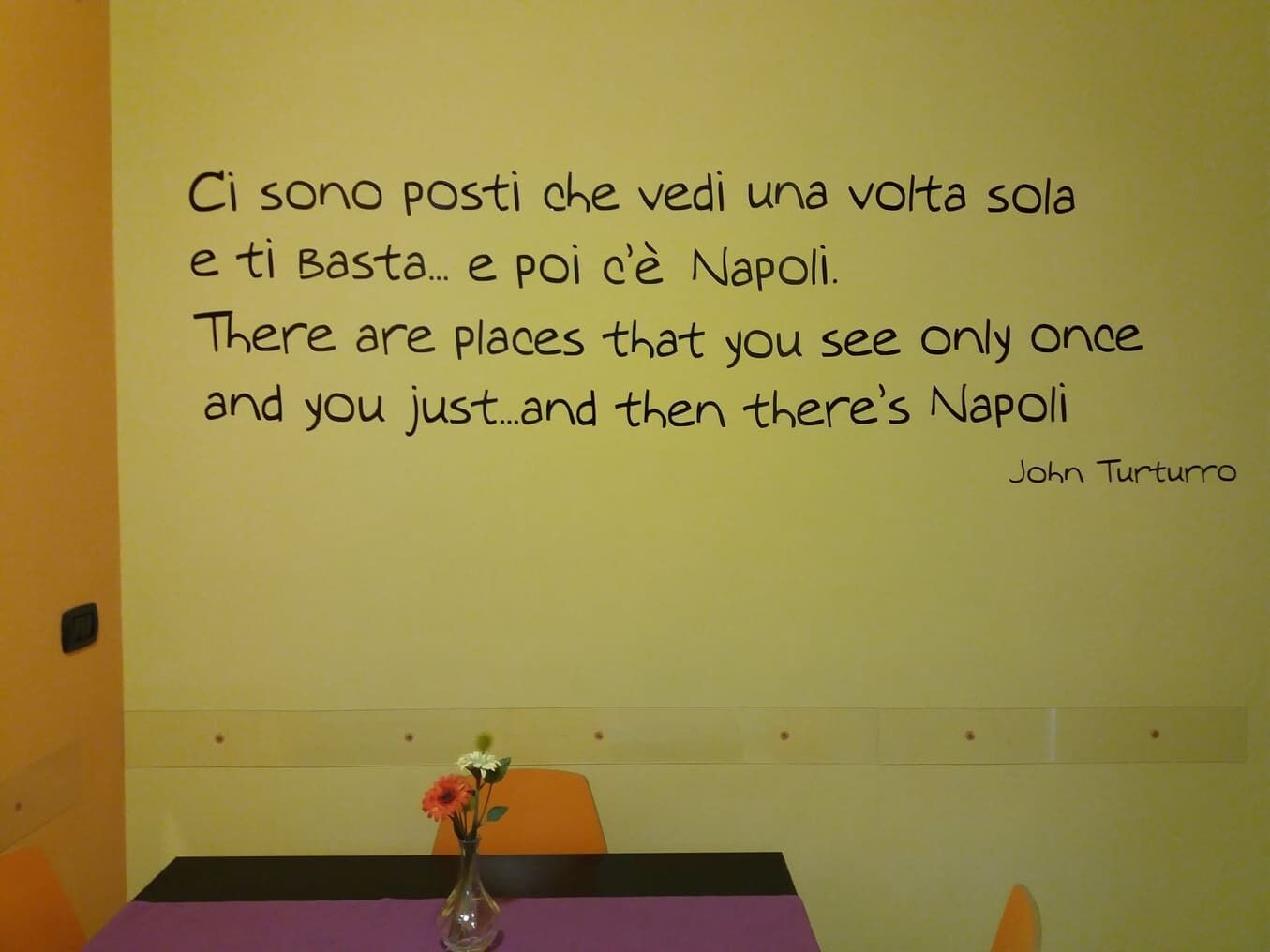 A quote on a wall in a room with a purple table and a vase of flowers.