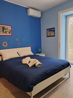 En seng med turkis sengetæppe og hvide puder, et natbord med lampe, samt en væg med dekorative detaljer.