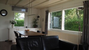 Ett modernt vardagsrum med en svart lädersoffa, ett matbord med stolar, en köksdel med ett vitt skåp och stora fönster som ger utsikt över grönområden utanför.