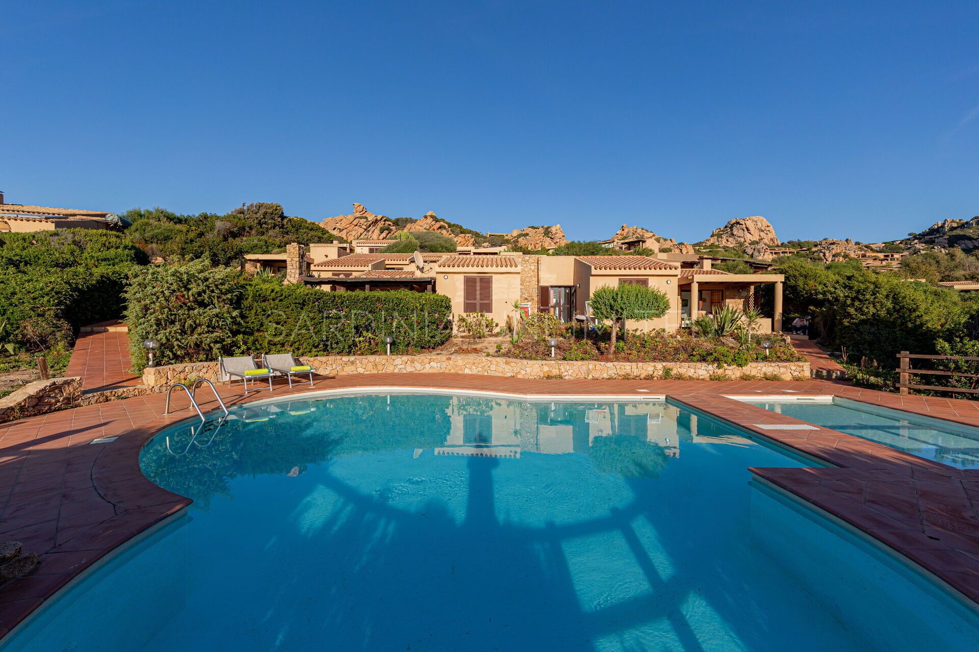 A pool area with a clear view of a building with multiple rooms, surrounded by greenery and rocky hills.