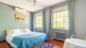 Une chambre à coucher avec un lit, un ventilateur de plafond, une fenêtre avec des rideaux, un radiateur et une petite table.