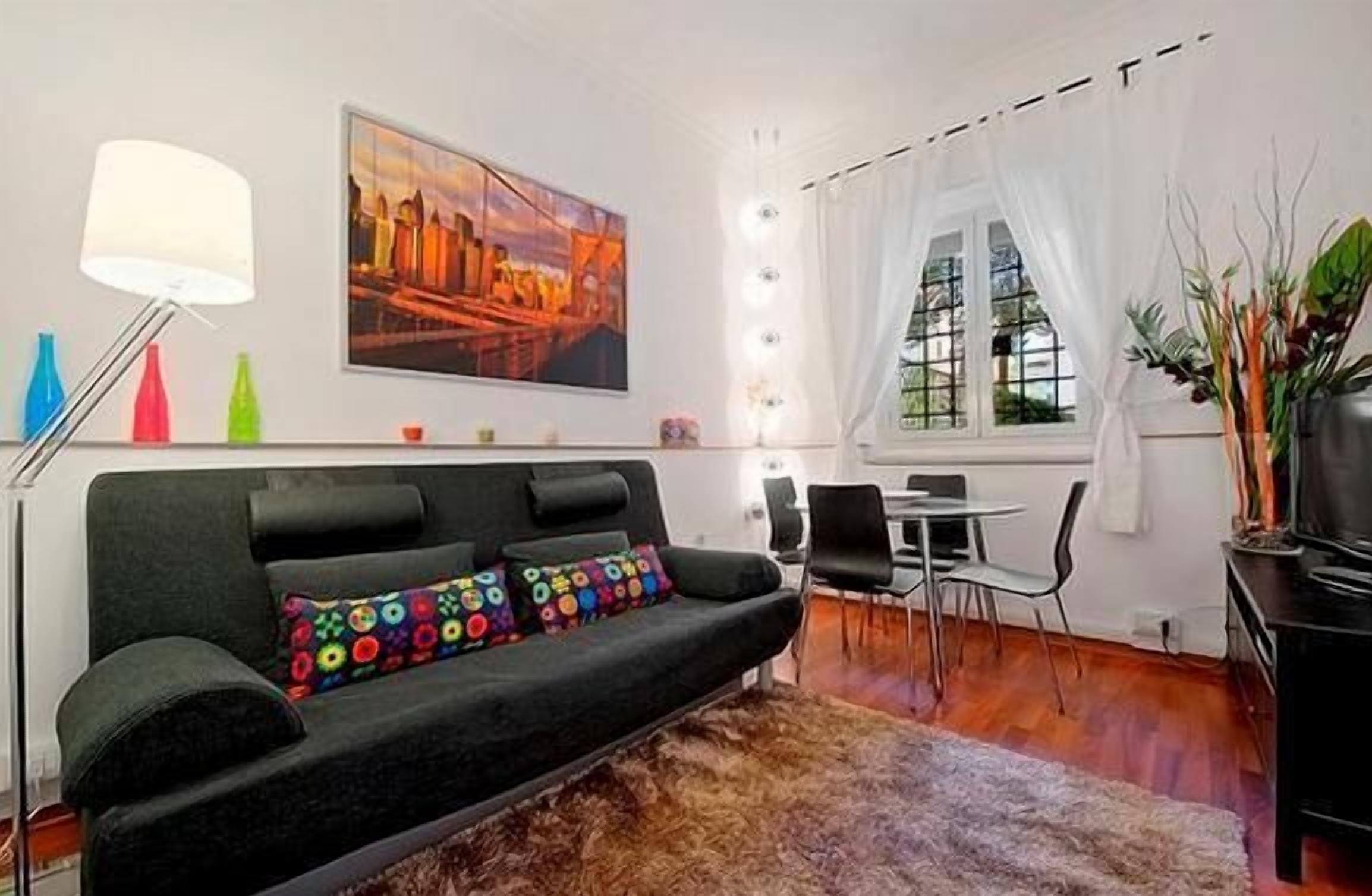Ein modernes Wohnzimmer mit einer schwarzen Couch, farbenfrohen Kissen, einem Gemälde an der Wand, einem Esstisch mit Stühlen und einem Fenster mit Vorhängen.
