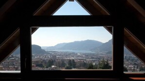 A view from a window with a triangular roof, overlooking a cityscape with mountains and a lake. - House / Villa - Lecco (Lecco)