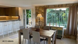 Un comedor con pared de piedra, una mesa con sillas, una lámpara y vistas a la vegetación por una ventana.