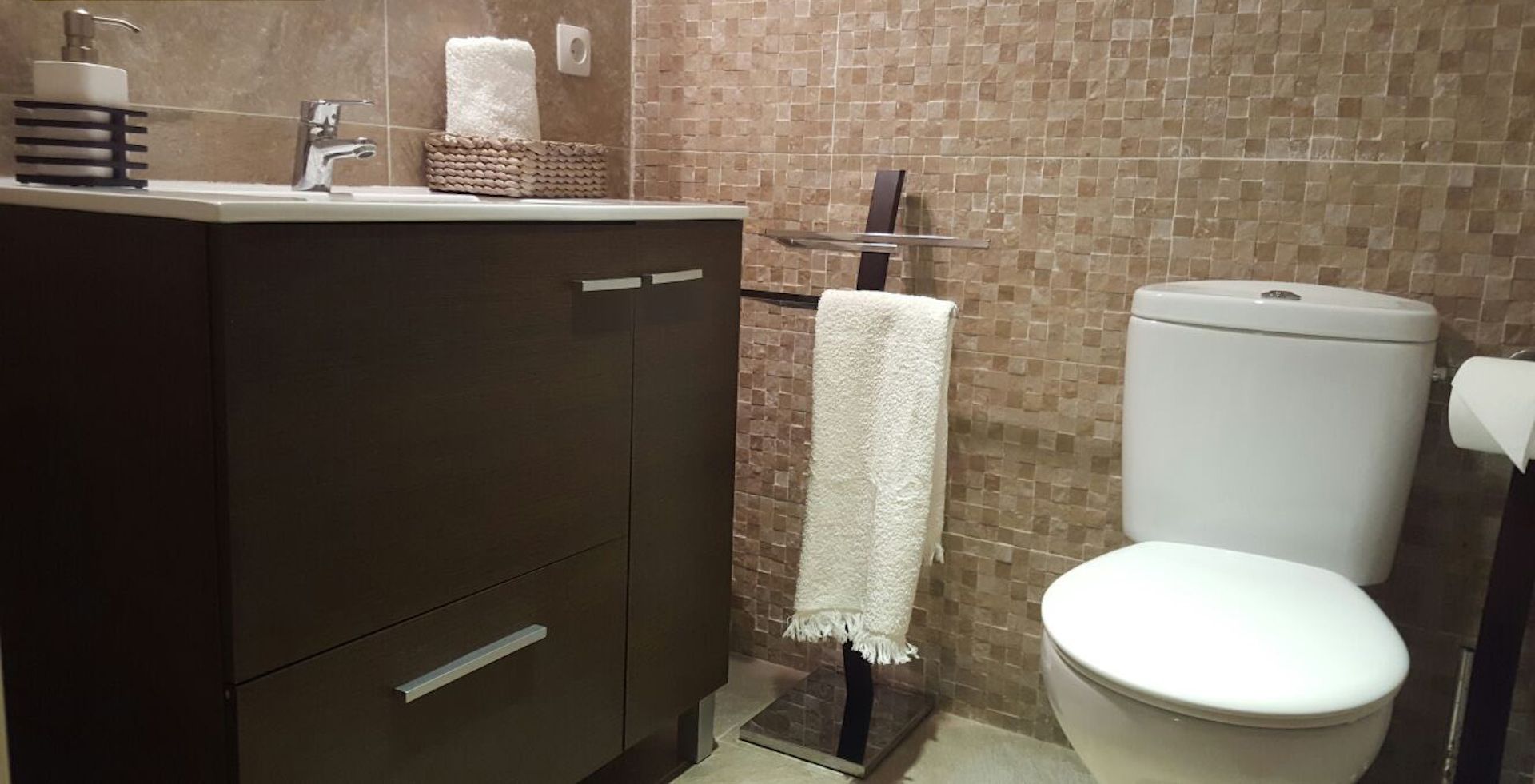 Une salle de bain avec un meuble-lavabo marron foncé, des toilettes blanches et un mur recouvert de carreaux.